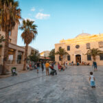 Gezellige sfeer op het Titou-plein in Heraklion, Kreta, met palmbomen, mensen die wandelen en de historische Sint-Tituskerk op de achtergrond.