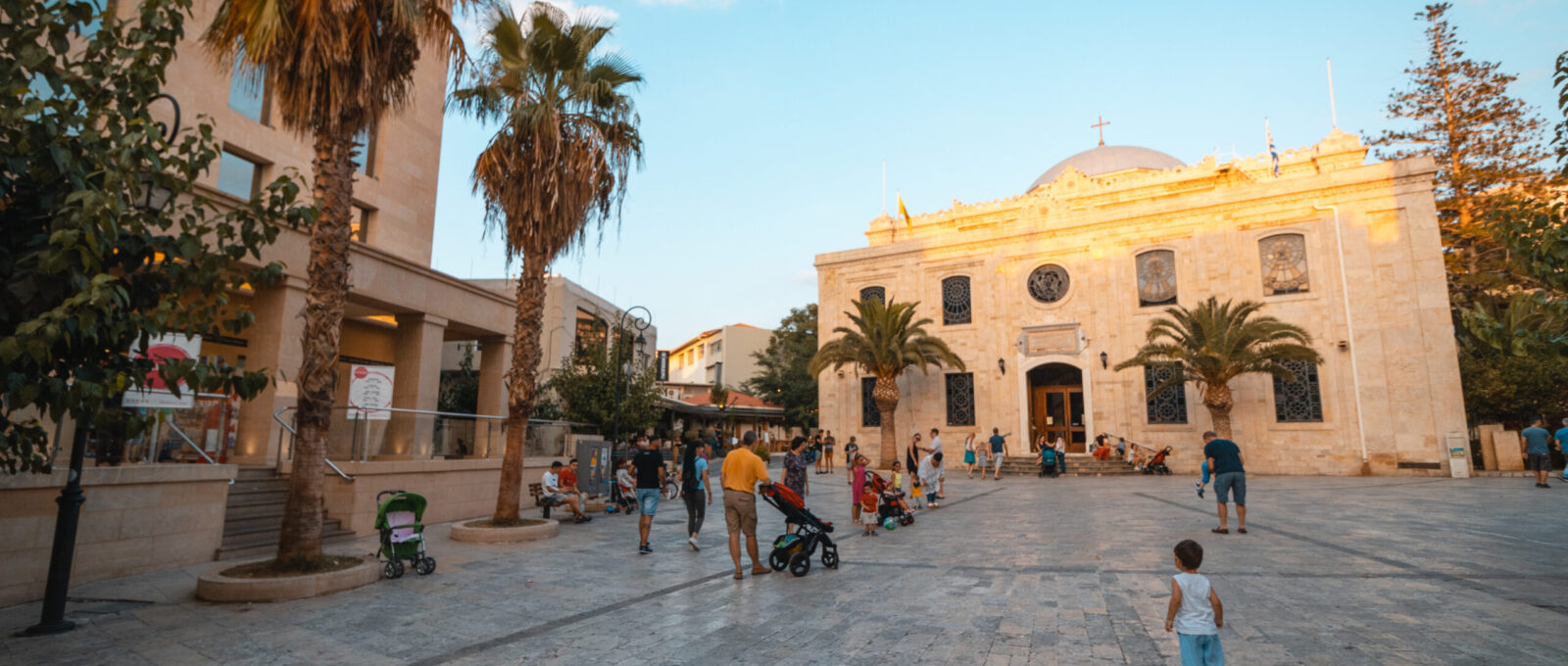 Gezellige sfeer op het Titou-plein in Heraklion, Kreta, met palmbomen, mensen die wandelen en de historische Sint-Tituskerk op de achtergrond.