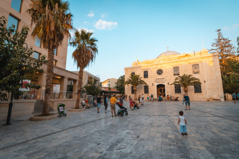 Gezellige sfeer op het Titou-plein in Heraklion, Kreta, met palmbomen, mensen die wandelen en de historische Sint-Tituskerk op de achtergrond.