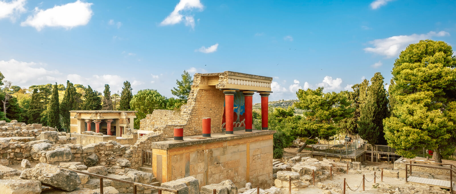 Ruïnes van het Paleis van Knossos op Kreta, met iconische rode zuilen en kleurrijke fresco’s, omringd door groene bomen en een blauwe lucht.