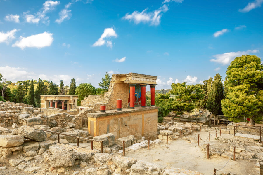 Ruïnes van het Paleis van Knossos op Kreta, met iconische rode zuilen en kleurrijke fresco’s, omringd door groene bomen en een blauwe lucht.