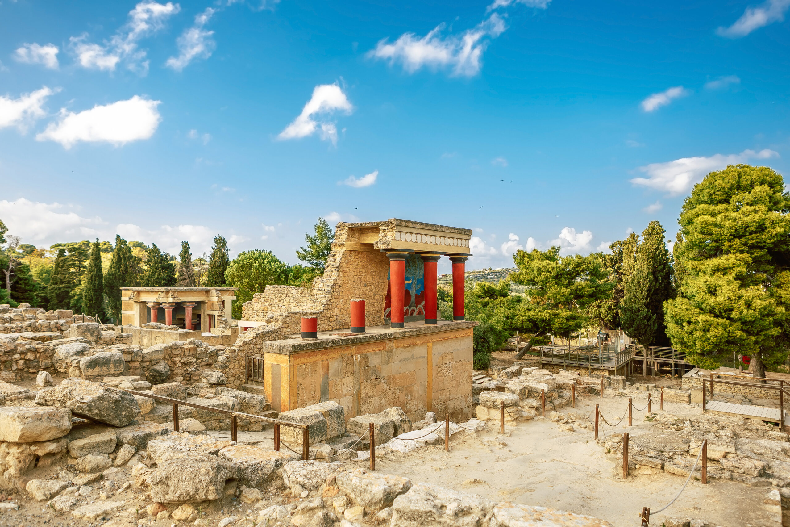 Ruïnes van het Paleis van Knossos op Kreta, met iconische rode zuilen en kleurrijke fresco’s, omringd door groene bomen en een blauwe lucht.