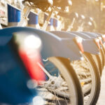 Een rij blauwe deelfietsen netjes op een rij bij een fietsenstalling, verlicht door warm zonlicht. De focus ligt op de achterwielen en spatborden, terwijl de achtergrond vervaagd is.