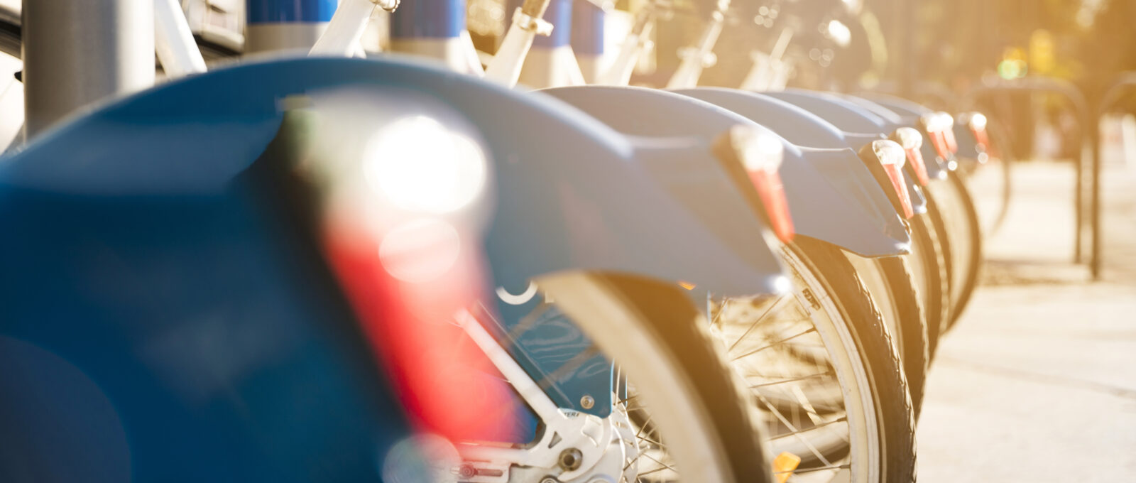 Een rij blauwe deelfietsen netjes op een rij bij een fietsenstalling, verlicht door warm zonlicht. De focus ligt op de achterwielen en spatborden, terwijl de achtergrond vervaagd is.