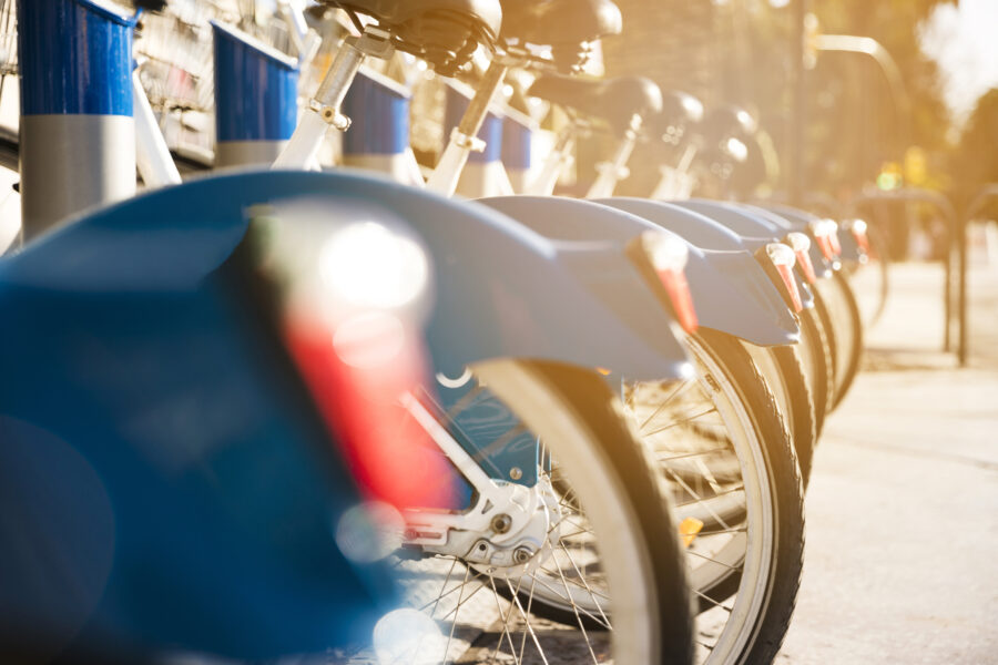 Een rij blauwe deelfietsen netjes op een rij bij een fietsenstalling, verlicht door warm zonlicht. De focus ligt op de achterwielen en spatborden, terwijl de achtergrond vervaagd is.