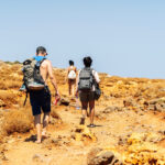 Drie wandelaars met rugzakken lopen over een droog, stenig pad onder een strakblauwe lucht op Kreta.