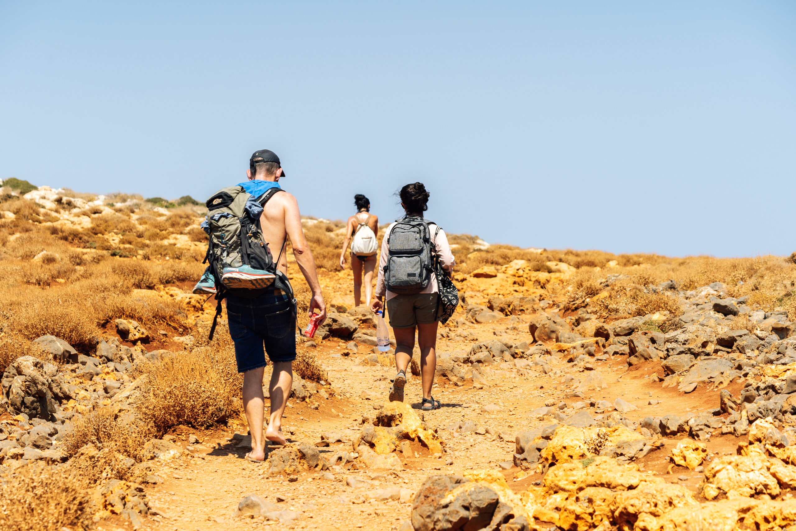 Drie wandelaars met rugzakken lopen over een droog, stenig pad onder een strakblauwe lucht op Kreta.